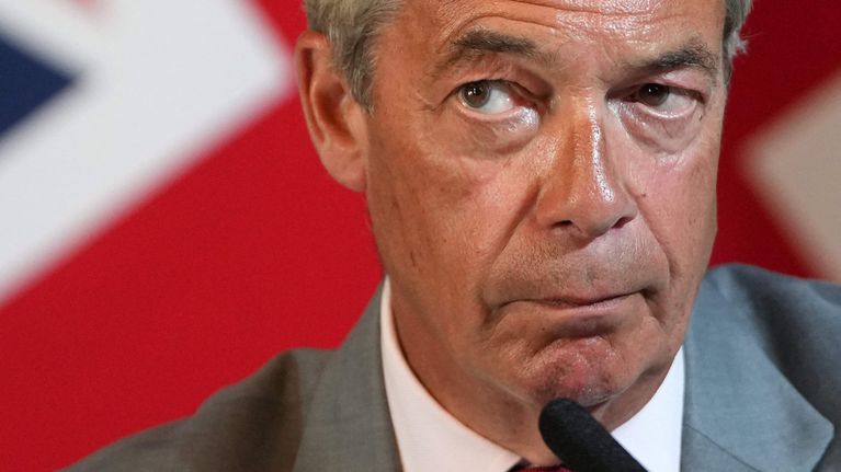 Brittipoliitikko Nigel Farage on lähikuvassa. Hän katsoo sivulle vakavailmeisenä. Hänen edessään on mikrofoni ja takanaan Britannian lippu.