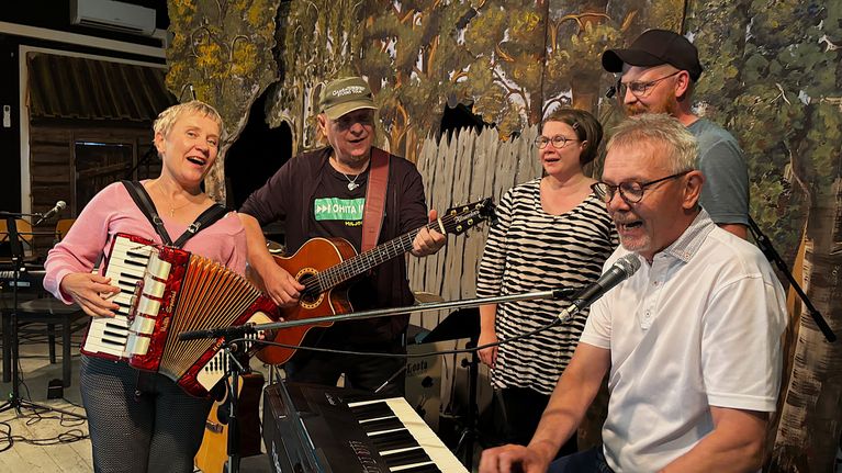 Viisi ihmistä musisoi näyttämöllä kosketinsoittajan ympärillä. Nainen soittaa haitaria ja lippalakkipäinen mies akustista kitaraa.