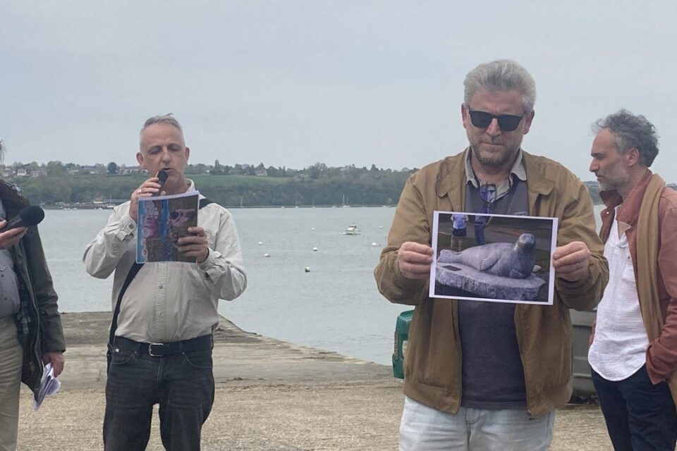 Dominique Bognet prend la parole pour évoquer le projet de statue, tandis qu’un intervenant montre une photo de la statue du phoque André, aux États-Unis.