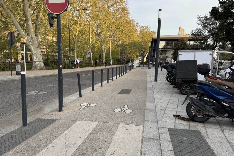 Un début de piste cyclable matérialisé au sol, qui s'arrête à l'entrée du parking de la gare, au fond de l'image 