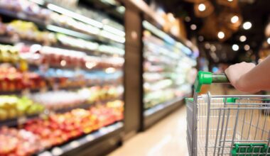 Woman Hand Hold Supermarket Shopping Cart With Abstract Blur Organic Fresh Fruits And Vegetable On Shelves In Grocery Store Defocused Bokeh Light Background