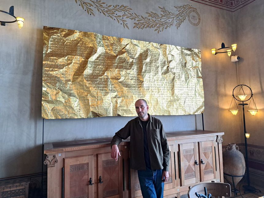 Gabriel Leger dans la bibliothèque de la Villa, devant l'oeuvre qui porte le nom de l'exposition : 'L'Or du temps'. Ce texte étant un assemblage de poèmes grecs, de l'Antiquité à nos jours. Laiton perforé et riveté, structure acier. 19 mai 2025
