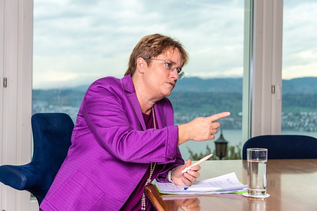 Magdalena Martullo-Blocher en discussion, vêtue d’une veste violette, assise à une table avec un verre d’eau et des documents à Herrliberg.