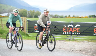 Adam Becket in a beige jacket rides along next to Katherine Moore in a