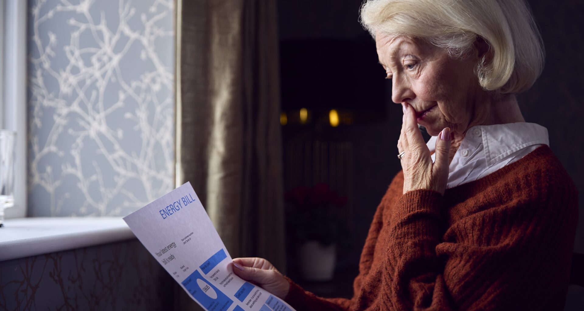 Senior Woman With Usa Energy Bill Trying To Keep Warm By Radiator In Cost Of Living Energy Crisis