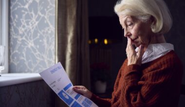 Senior Woman With Usa Energy Bill Trying To Keep Warm By Radiator In Cost Of Living Energy Crisis