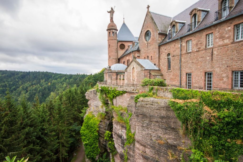 Château avec tours en pierre perché sur une falaise abrupte entourée d'une forêt verte dense sous un ciel nuageux.