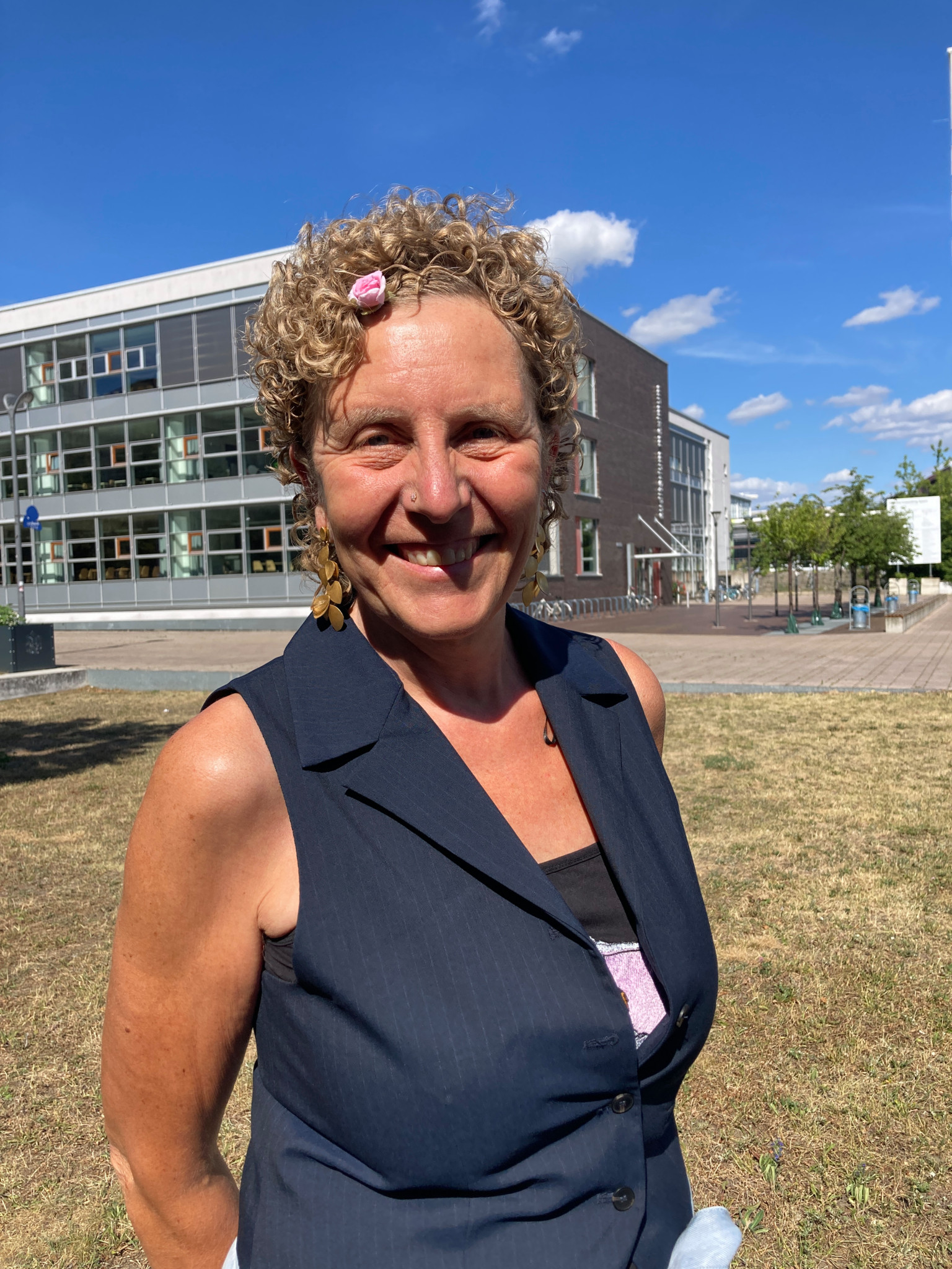 Femme souriante aux cheveux bouclés, portant un gilet noir, se tenant sur une pelouse devant un bâtiment moderne avec un ciel bleu en arrière-plan.