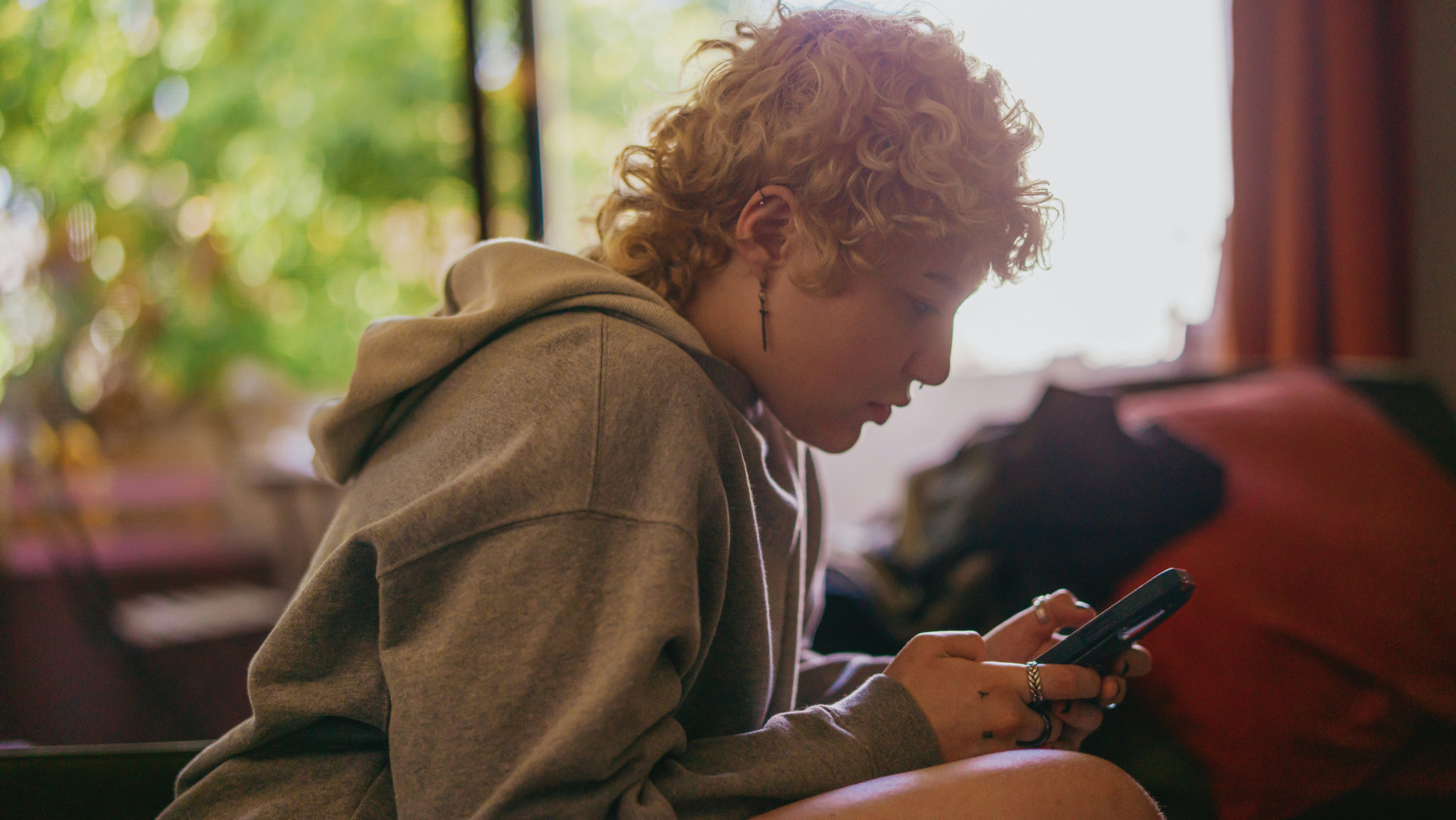 Jeune personne aux cheveux bouclés, vêtue d’un sweat à capuche gris, utilisant un téléphone portable dans une pièce lumineuse.