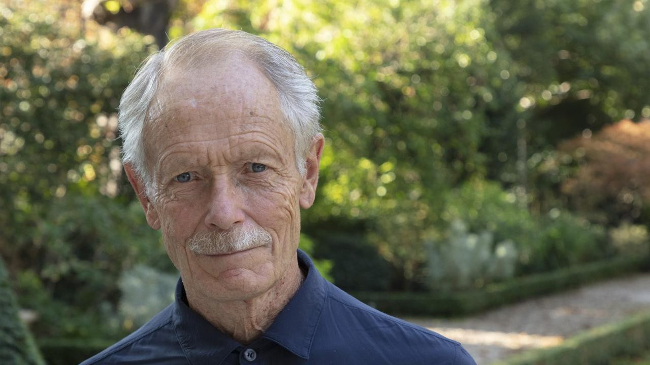 Homme aux cheveux gris, moustache, vêtu d’une chemise bleue, se tenant dans un jardin verdoyant.
