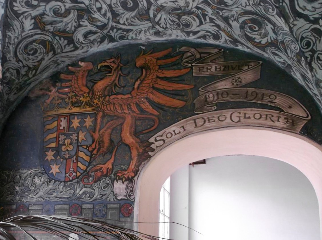 Une fresque murale avec un aigle héraldique tenant un blason richement décoré, entouré de motifs floraux et d’une bannière portant les dates 1910-1912 et le texte ’Soli Deo Gloria’.