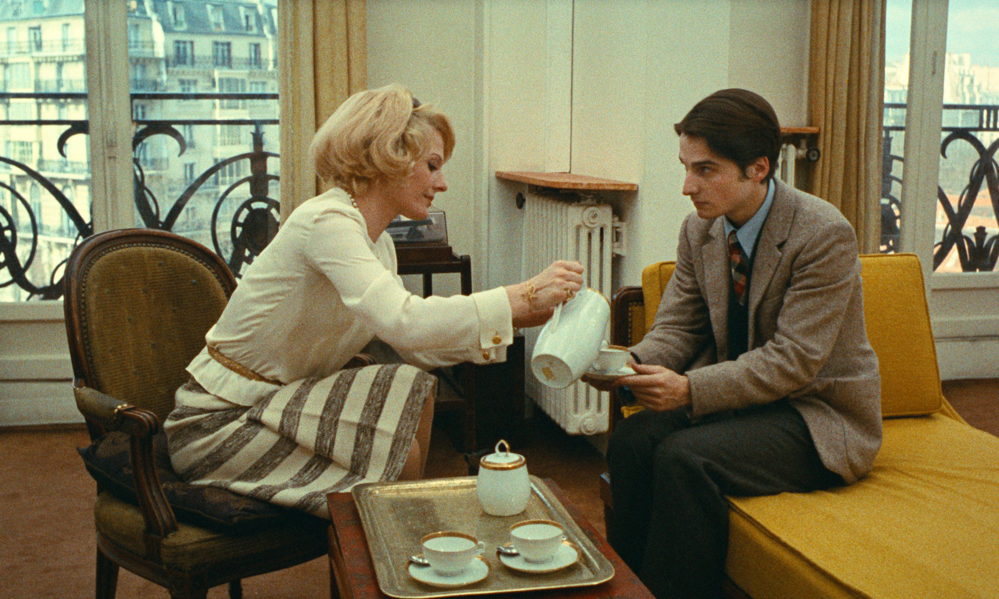 Une femme verse du thé dans la tasse d’un homme assis sur un canapé jaune, dans un salon avec vue sur des immeubles parisiens.