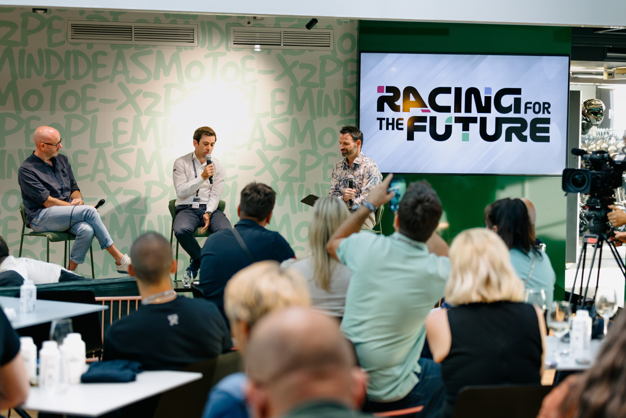 Trois hommes discutent sur scène lors d’un événement intitulé ’Racing for the Future’, avec un public attentif.