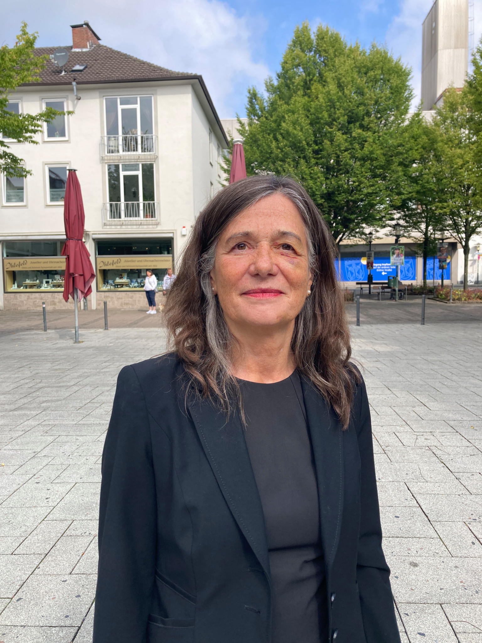 Femme aux cheveux bruns portant un blazer noir, debout sur une place pavée avec des arbres et des bâtiments en arrière-plan.