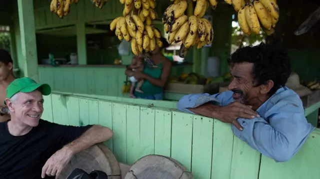 Dom sourit en parlant à un homme appuyé sur le rebord d'un balcon, avec des bananes qui pendent au-dessus.