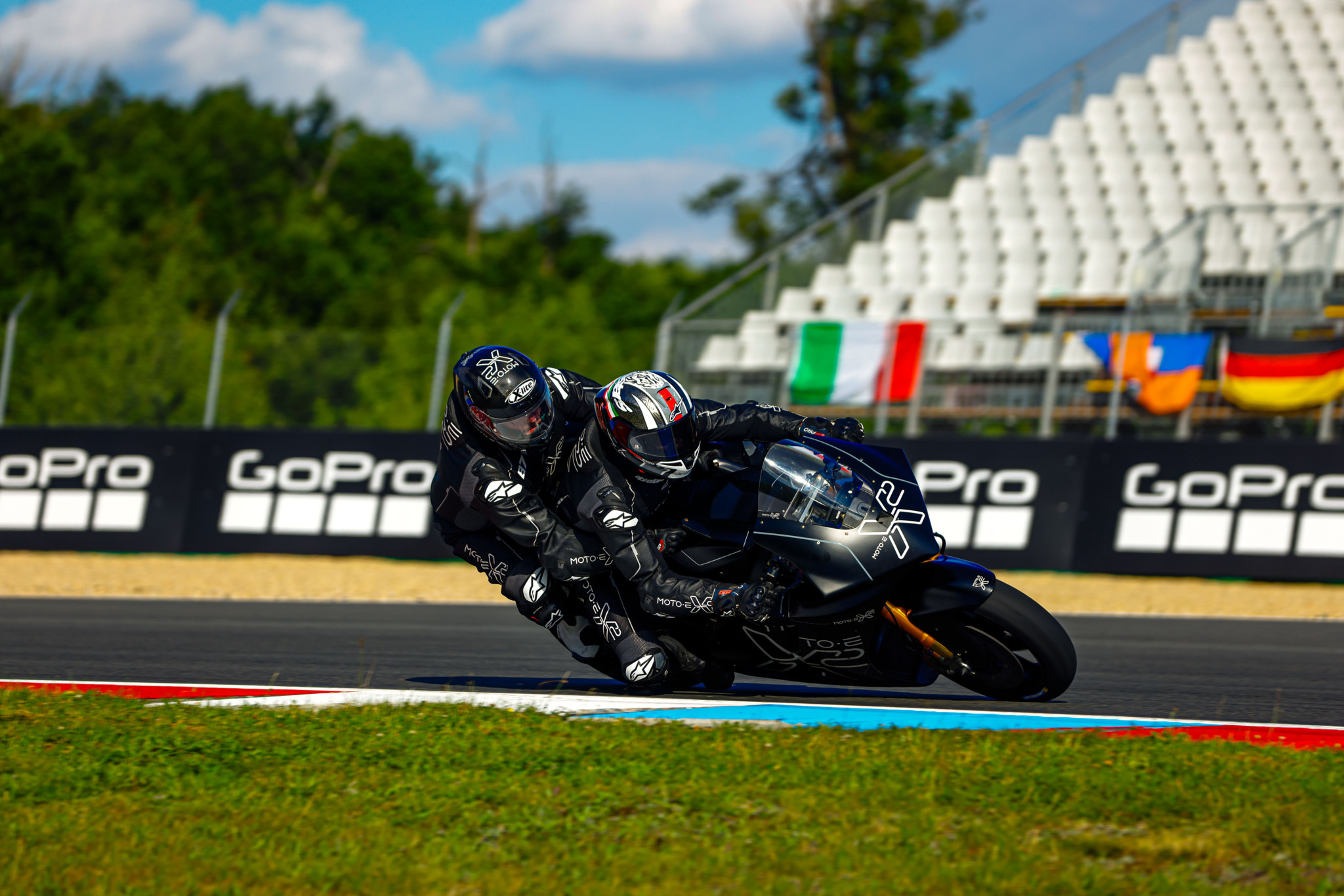 Deux motocyclistes en tenue noire prennent un virage serré sur un circuit de course, avec des tribunes et des drapeaux en arrière-plan.