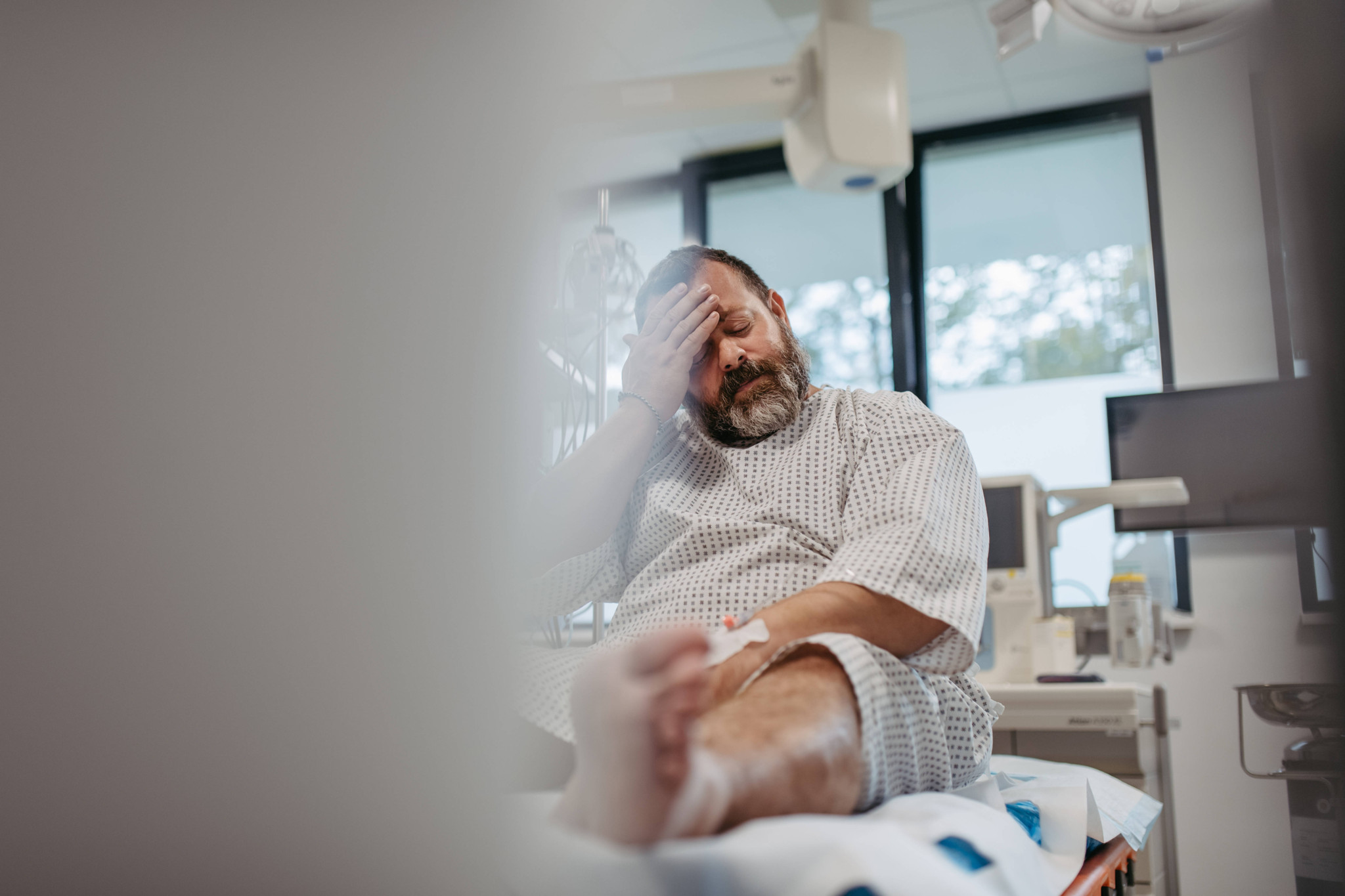 Patient en surpoids portant une blouse d’hôpital, assis dans une salle d’examen médical, tenant sa tête avec une expression de douleur et d’anxiété.