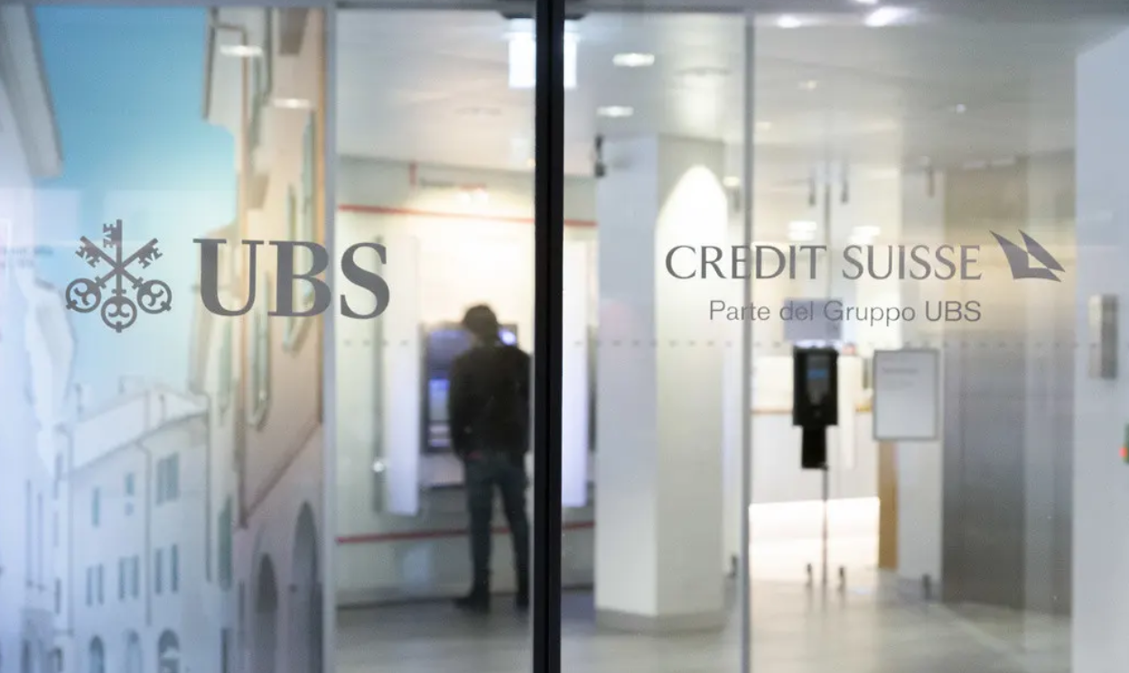 Entrée vitrée de la banque avec logos UBS et Credit Suisse, personne utilisant un distributeur automatique à l’intérieur.