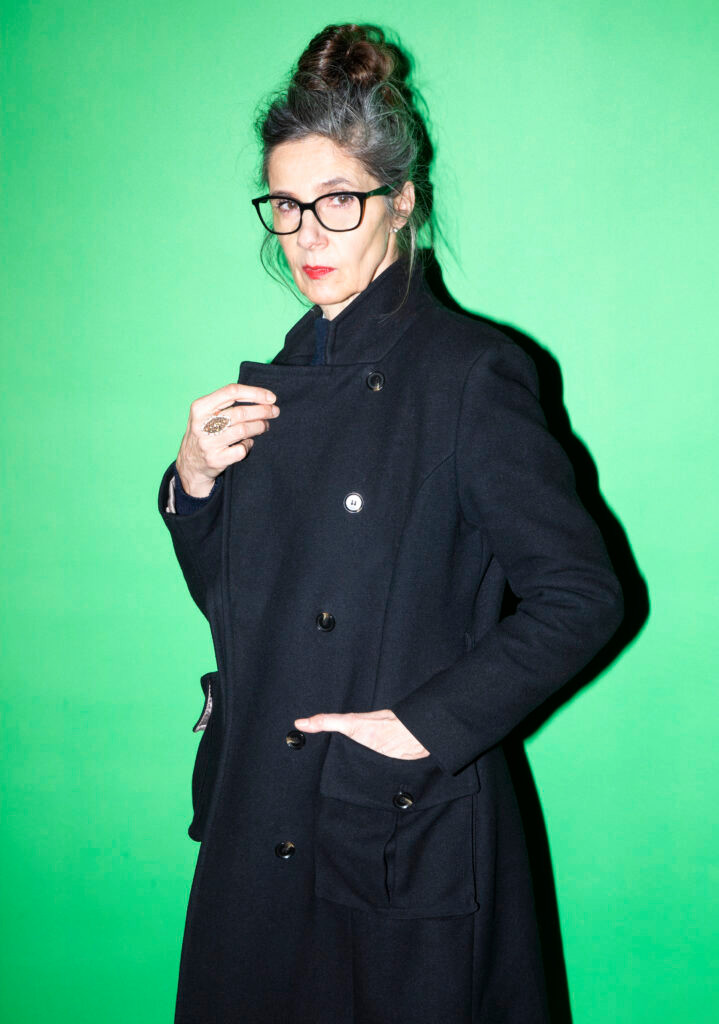 Femme aux cheveux attachés portant un manteau noir sur fond vert avec des lunettes.