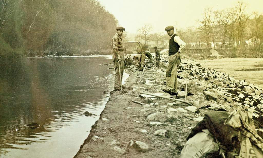 Deux hommes discutent au bord d’une rivière avec des vêtements vintage, pendant des travaux ou une activité de pêche.