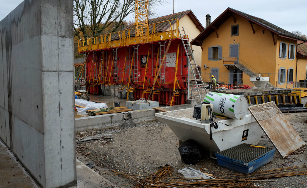 Chantier de construction avec échafaudages et matériaux, à côté de maisons jaunâtres. Ciel nuageux en arrière-plan.
