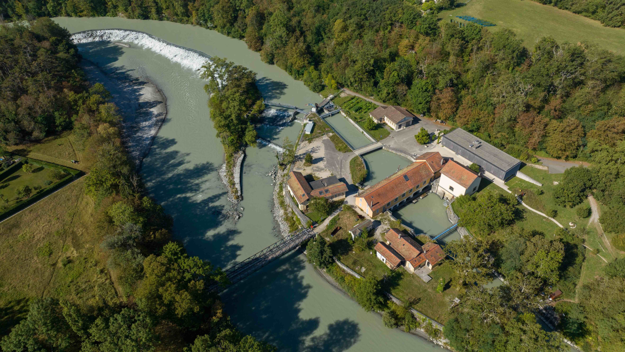 Vue aérienne d’un moulin à eau entouré de forêt verdoyante le long d’une rivière sinueuse.