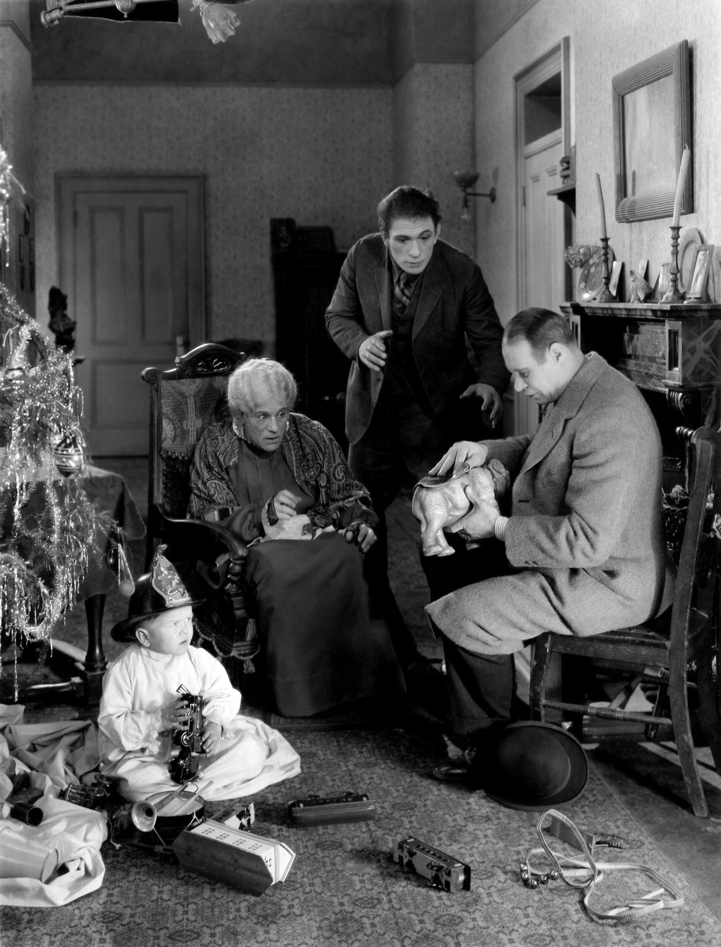 Scène de Noël en noir et blanc avec un bébé jouant au sol, une vieille femme assise avec un chat, deux hommes regardant un cadeau.