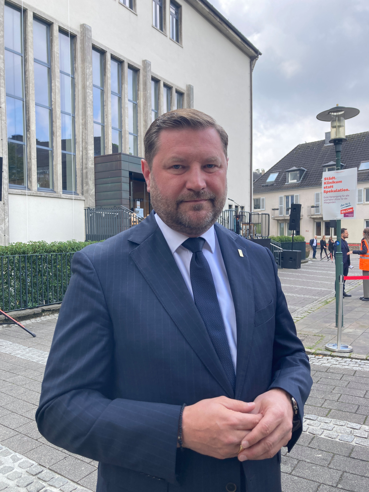 Un homme en costume sombre se tient devant un bâtiment moderne avec de grandes fenêtres.