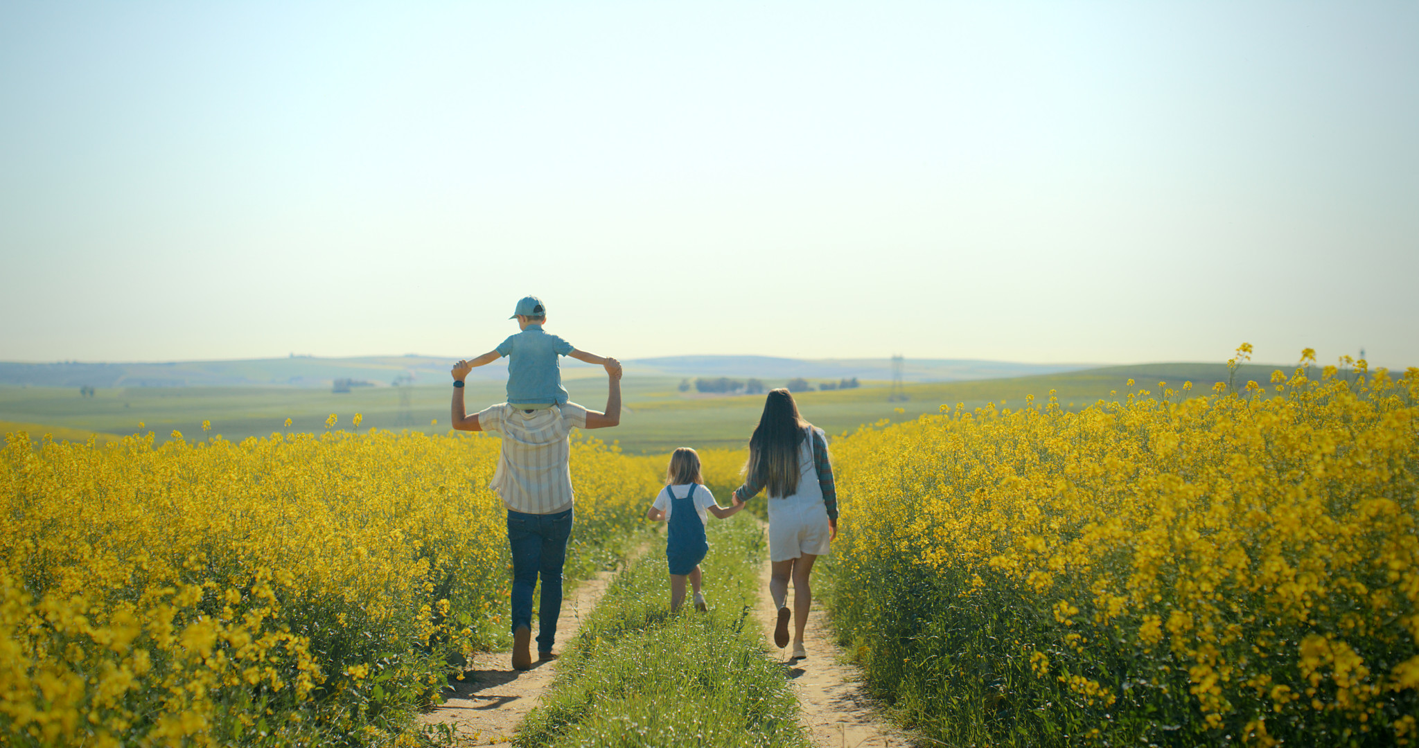 Famille heureuse se promenant dans un champ de fleurs jaunes sous un ciel bleu. Enfant sur les épaules du père, marchant avec la mère dans la campagne printanière.