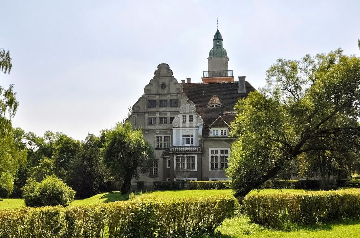 Grand bâtiment historique avec une architecture ancienne, entouré de verdure et d’arbres sous un ciel dégagé.