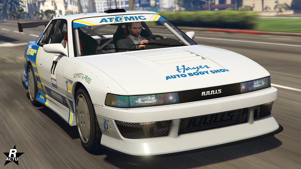 Cette image montre une voiture de sport blanche, l’Annis Remus, avec des détails en bleu et jaune, en mouvement dans le jeu GTA Online. La voiture porte plusieurs logos et inscriptions, dont ‘ATOMIC’ sur le pare-brise et le côté de la voiture, ‘Hayes Auto Body Shop’ sur le capot, et ‘REMUS’ en lettres noires sur la partie avant de la voiture. Le véhicule a un design sportif avec un aileron arrière, des jantes en alliage et un pare-chocs avant modifié pour un look plus agressif. Un personnage du jeu avec une tête floutée conduit la voiture; l’intérieur noir de la voiture est partiellement visible. À l’arrière-plan, on peut voir des bâtiments flous et des palmiers typiques de Los Santos.