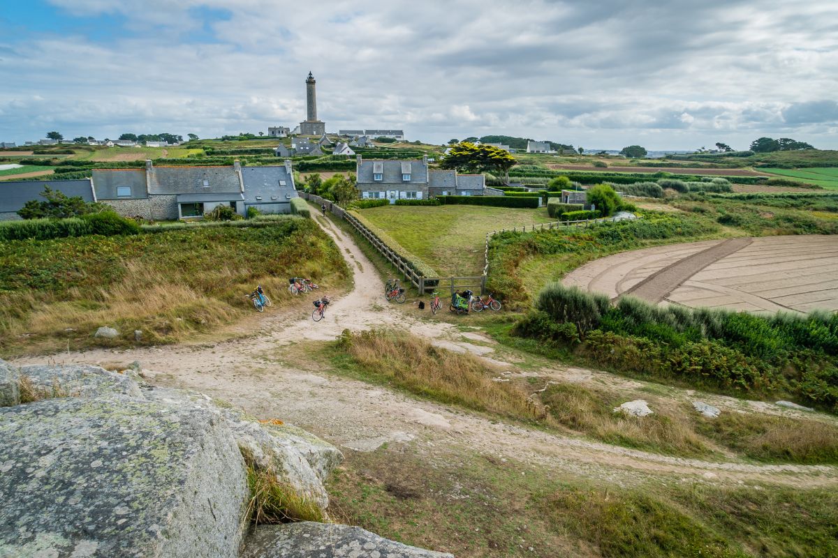 Un paysage rural avec un phare en arrière-plan, des maisons, des champs et un sentier menant à travers un terrain herbeux. Des vélos sont garés près du chemin et des arbres parsèment l’horizon sous un ciel nuageux.