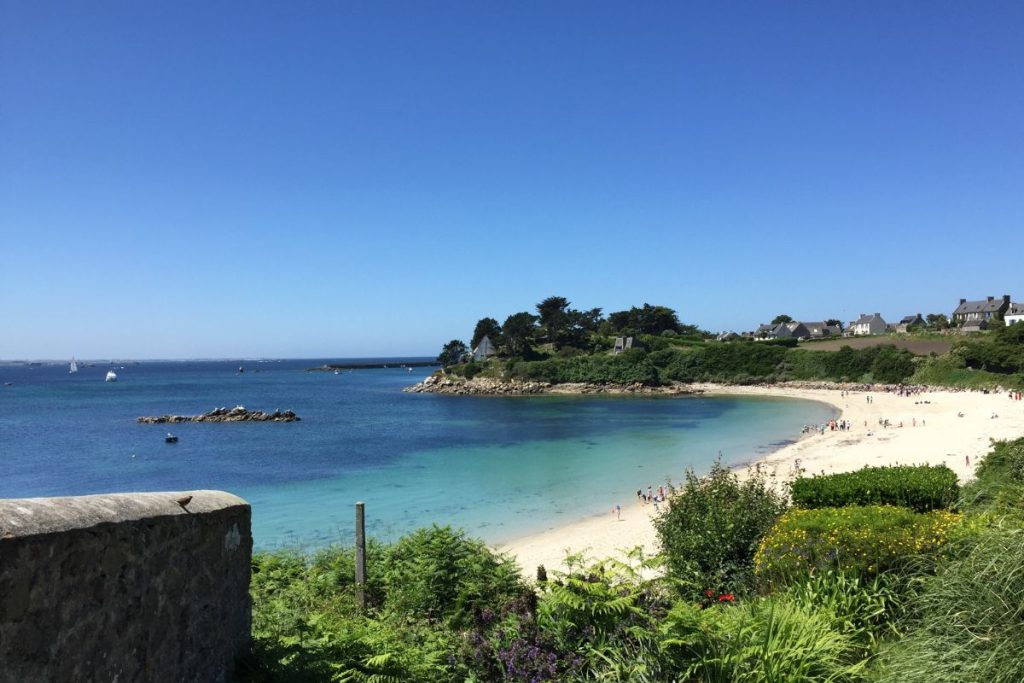 Une scène de plage sereine avec un ciel bleu clair, des eaux turquoise tranquilles et un rivage sablonneux bordé de végétation verte et quelques personnes, quelques maisons visibles sur le côté droit.