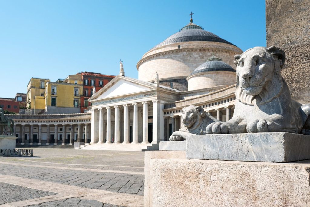 L'église Saint-François-de-Paule avec ses dômes et ses colonnes sur la Piazza del Plebiscito à Naples, avec une statue de lion en pierre au premier plan et des bâtiments colorés à l'arrière-plan.
