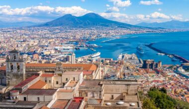 Vue panoramique de Naples, Italie, avec les bâtiments de la ville au premier plan, la baie et le mont Vésuve à l'arrière-plan, sous un ciel partiellement nuageux.