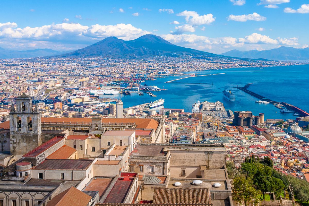 Vue panoramique de Naples, Italie, avec les bâtiments de la ville au premier plan, la baie et le mont Vésuve à l'arrière-plan, sous un ciel partiellement nuageux.