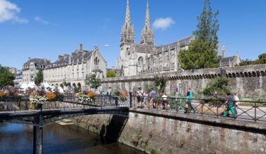 Vue ensoleillée sur une cathédrale gothique historique à côté d'une rivière avec des gens traversant un pont orné de fleurs.