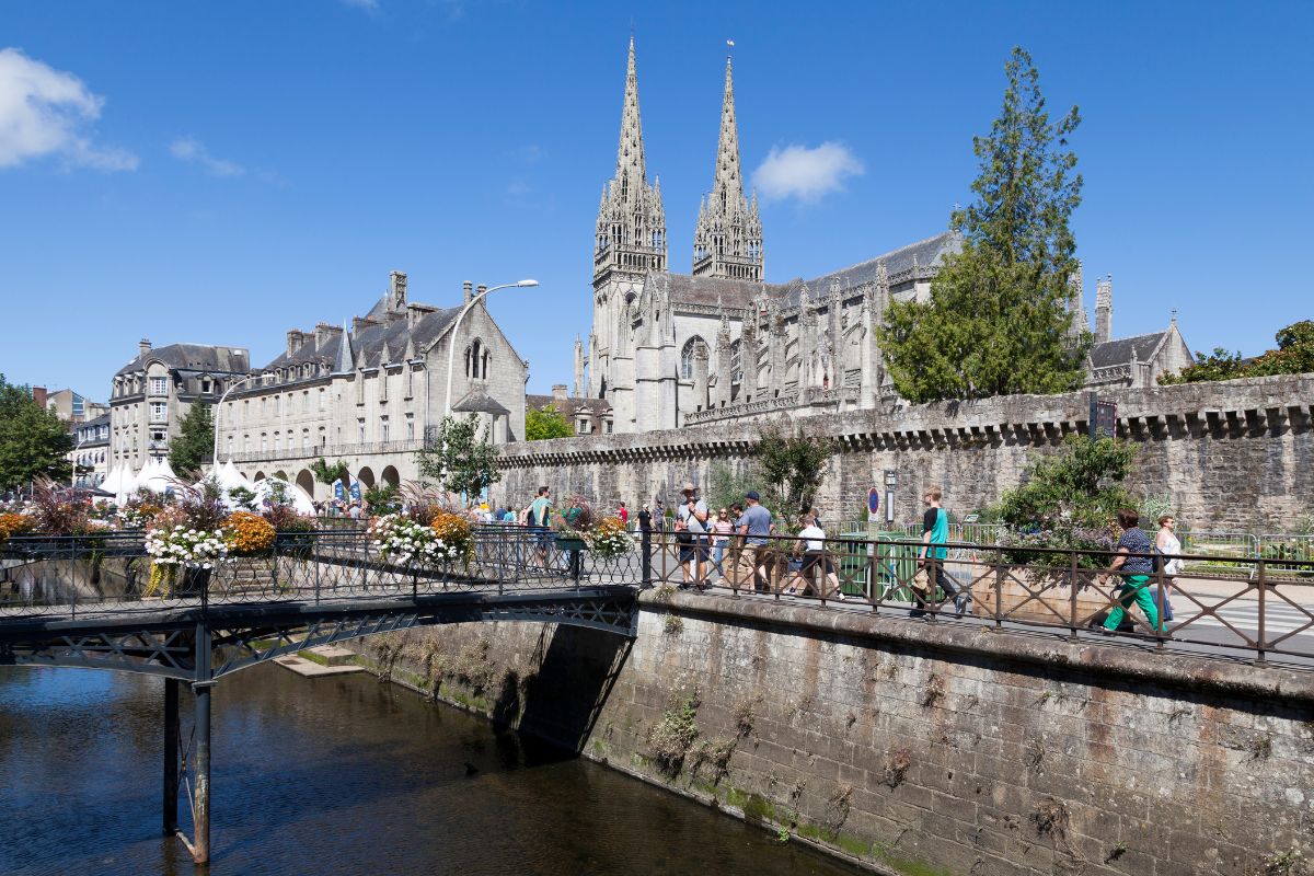 Vue ensoleillée sur une cathédrale gothique historique à côté d'une rivière avec des gens traversant un pont orné de fleurs.