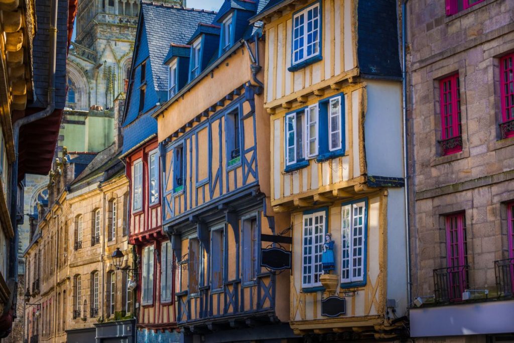 Ruelle pittoresque et étroite bordée de maisons à colombages colorées dans une ville européenne, avec un aperçu de la flèche de la cathédrale en arrière-plan.