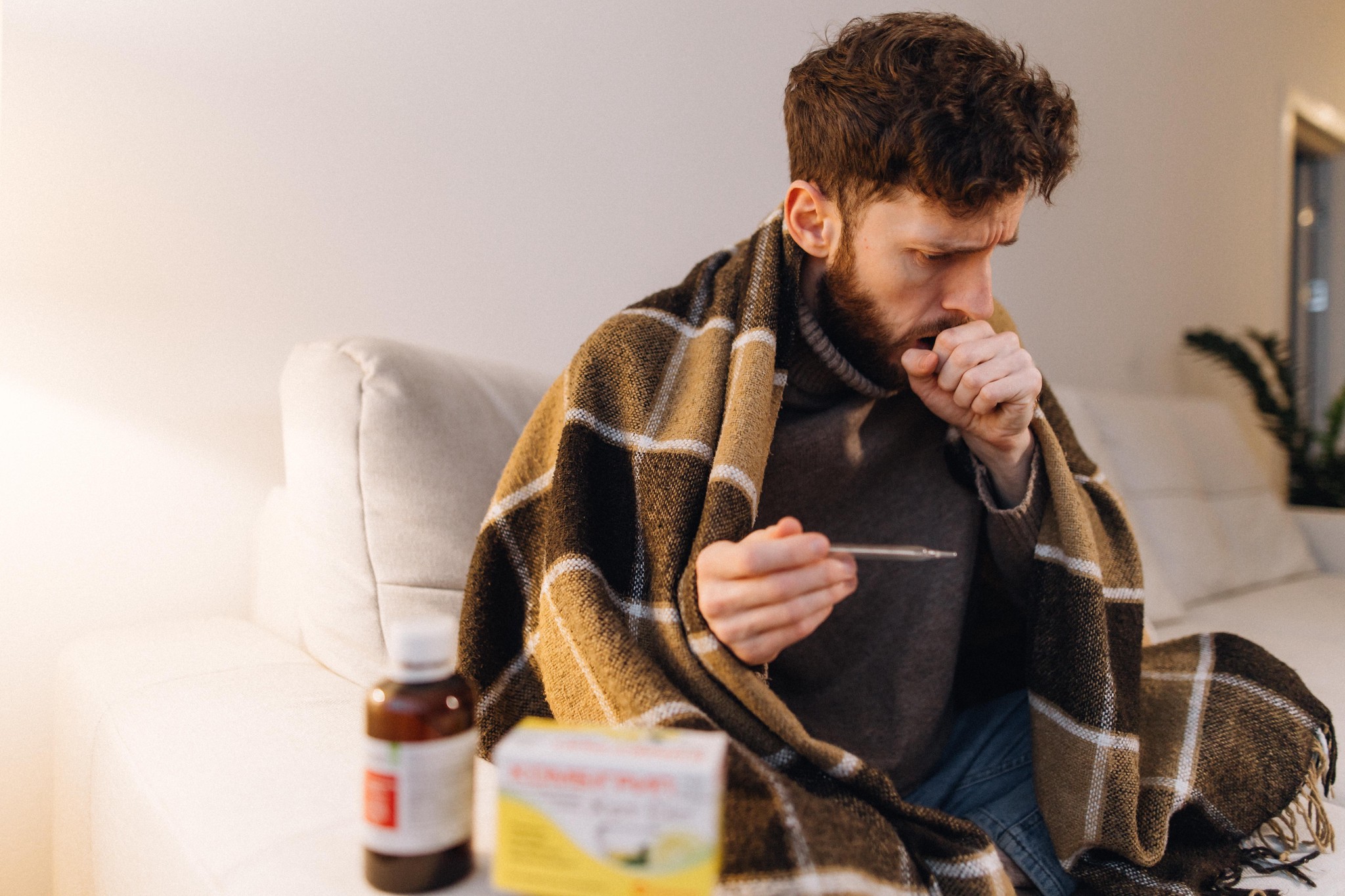 Un homme, enveloppé dans une couverture, mesure sa température et tousse, illustrant une approche proactive des soins à domicile pendant une maladie.