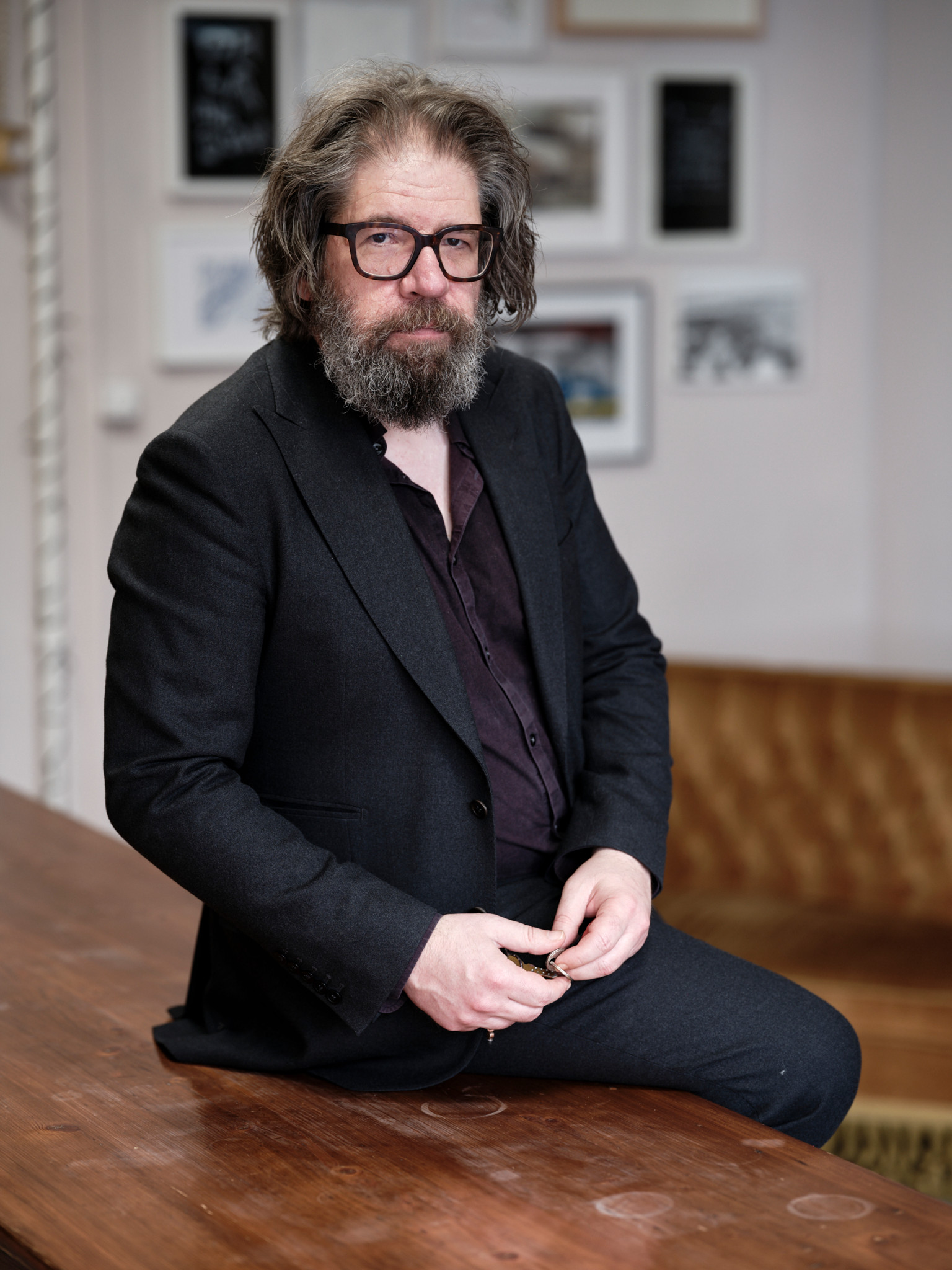 Homme barbu avec lunettes et costume sombre assis sur une table en bois, intérieur avec cadres au mur.