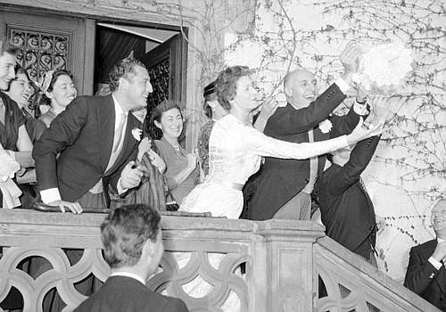 Mariage avec mariée en robe blanche jetant son bouquet depuis un balcon, entourée de personnes souriantes.