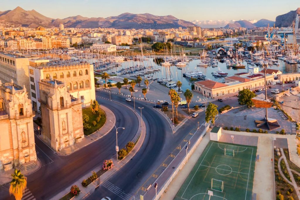 Vue aérienne d'un port de plaisance avec de nombreux bateaux amarrés, de bâtiments urbains adjacents, d'un terrain de sport, de routes sinueuses et de montagnes lointaines sous un ciel dégagé.