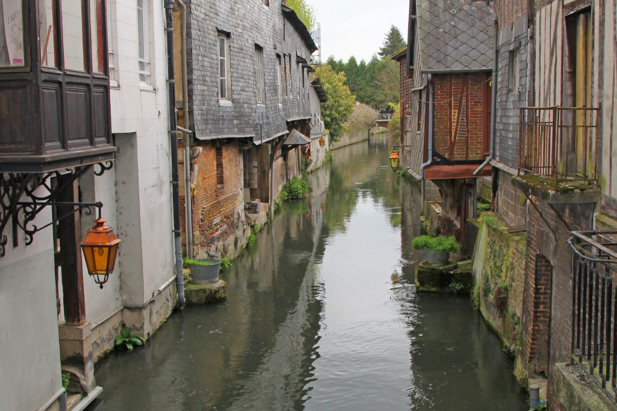 Un canal étroit coule entre de vieux bâtiments avec des lanternes et de la verdure, créant une scène pittoresque.