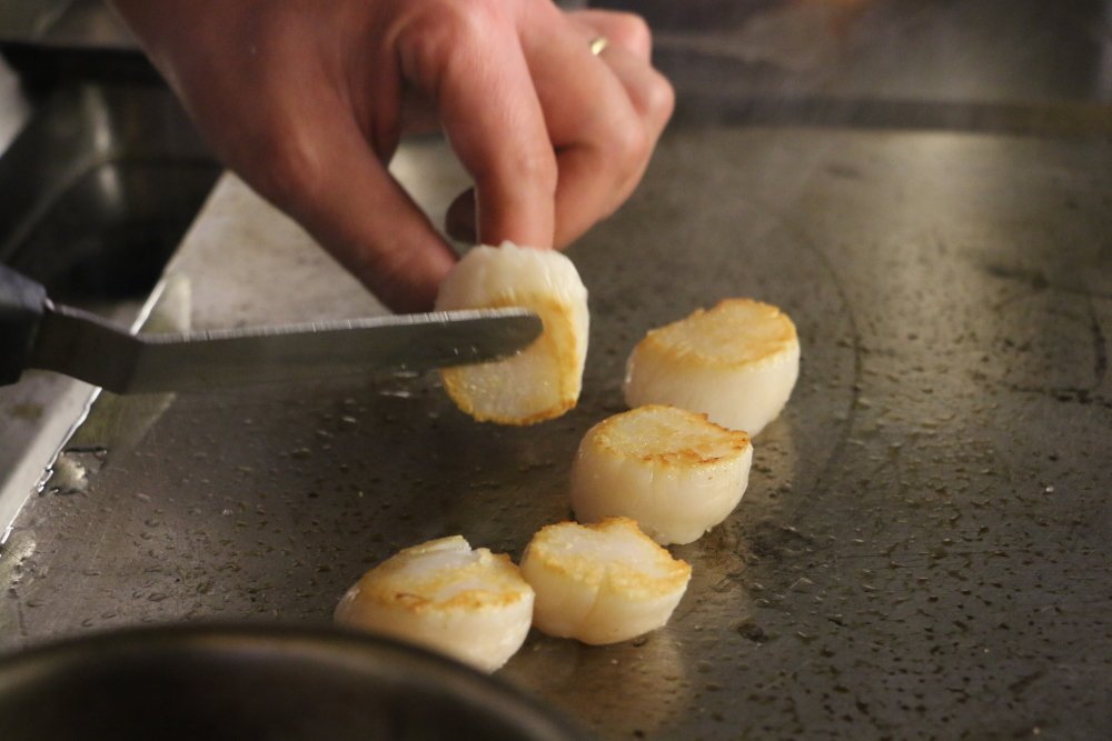 Les Saint-Jacques sont cuites directement sur une plancha préalablement huilée. Il faut qu'une croûte extérieure se forme pour les retourner. A la fin de la cuisson, pour apporter de la gourmandise, on met une noix de beurre.