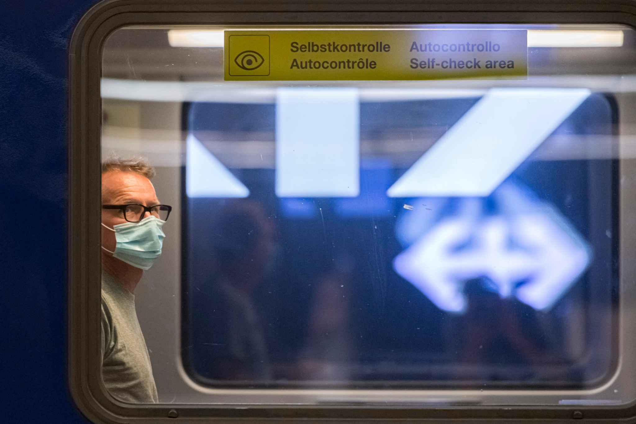 Un homme portant un masque chirurgical sort d’un train à la gare principale, sous un panneau indiquant ’Autocontrôle’ en trois langues.
