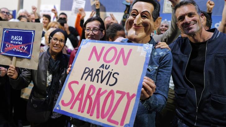 Un habitant de la région, portant un masque à l’effigie de Nicolas Sarkozy, brandit une pancarte pour protester contre la proposition du maire de Nice de donner à la place située devant le futur commissariat le nom de l’ancien président français Nicolas Sarkozy, à Nice, le 21 octobre 2025. 