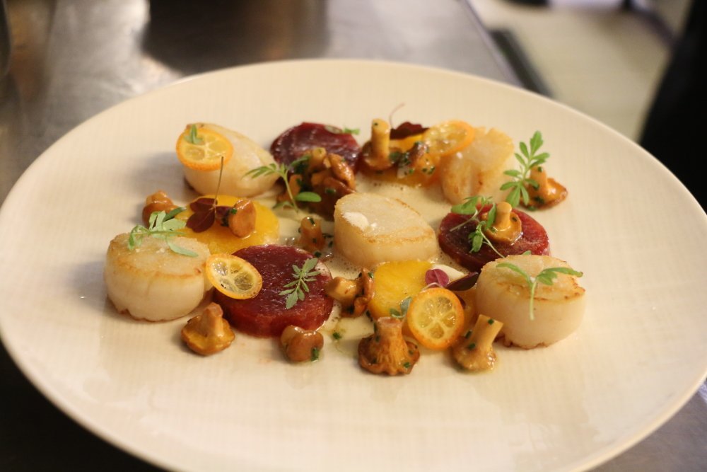 Place au dressage du plat, une étape qui demande de la précision. Il faut disposer les Saint-Jacques dans l'assiette puis les lamelles de kumquat, les girolles et ensuite parsemer d'herbes aromatiques.