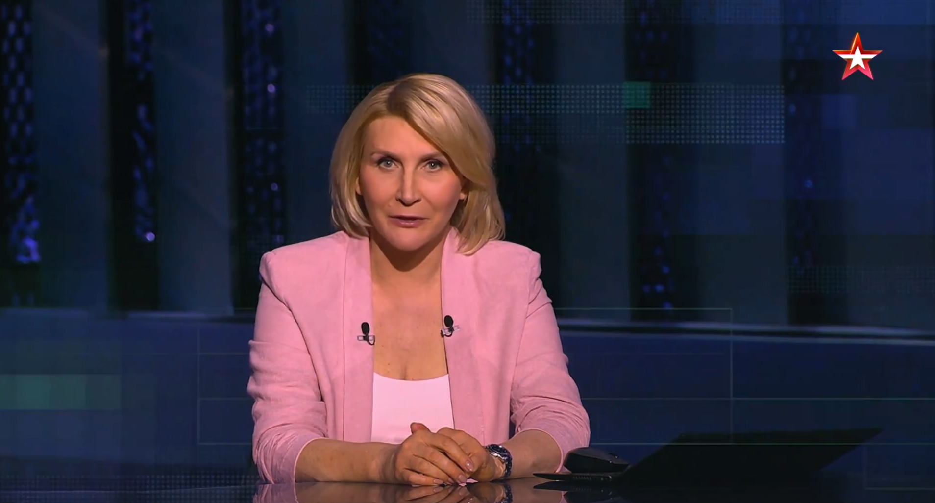 Présentatrice portant un blazer rose lors d’une émission satire russe générée par IA, Politukladchik, avec décor de studio moderne.