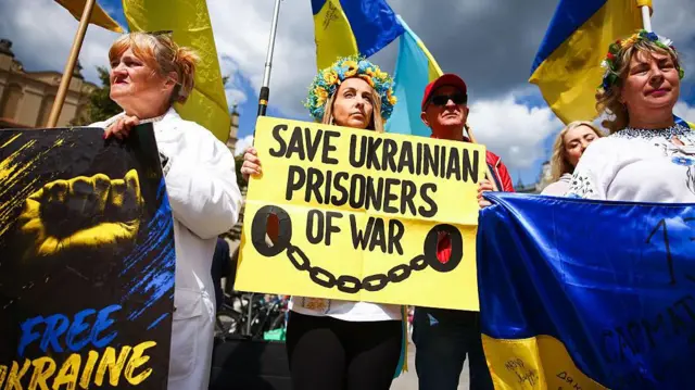Les manifestants photographiés dans un ciel essentiellement bleu tiennent des pancartes demandant la libération des prisonniers de guerre ukrainiens. Ils sont drapés dans les couleurs nationales de l'Ukraine et tiennent également une grande pancarte disant « Ukraine libre ».